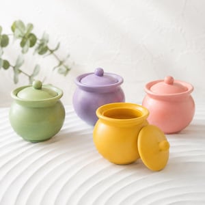 May include: Four small, pastel-colored ceramic jars with lids. The jars are in shades of green, purple, pink, and yellow. One yellow jar has its lid off, revealing the interior. The jars are arranged on a white, textured surface.
