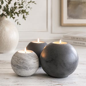 May include: Three spherical candle holders in varying sizes. The candle holders are gray and white, with a marble-like pattern on the smallest one. Each holder contains a lit candle, casting a warm glow. The arrangement is on a white wooden surface.