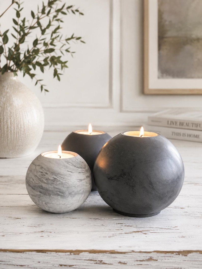 May include: Three spherical candle holders in varying sizes. The candle holders are gray and white, with a marble-like pattern on the smallest one. Each holder contains a lit candle, casting a warm glow. The arrangement is on a white wooden surface.