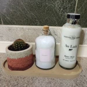 May include: A small cactus in a textured pink and grey pot, a glass bottle filled with white bath salts labelled Peony with a cork stopper, and a glass bottle of Scottish Fine Soaps Au Lait body milk, all arranged on an oval beige tray.