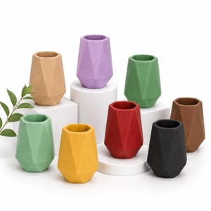 May include: A collection of eight geometric planters in various colors including tan, lavender, green, red, yellow, black, and brown. Each planter features a faceted, multi-sided exterior surface and an open top, arranged on white display pedestals.