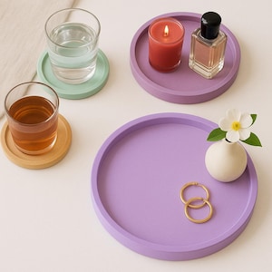 May include: Four colorful coasters with a purple, green, and yellow design. A clear glass with a beverage, a candle, and a bottle of perfume are on top of the coasters.