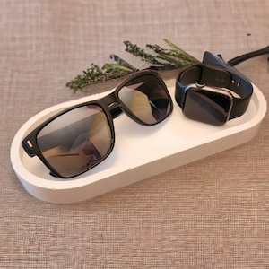 May include: A pair of black sunglasses with reflective lenses and a black smartwatch with a silicone strap resting on a white oval tray. The tray has a smooth, matte finish and a raised edge, providing a clean surface for these two accessories.