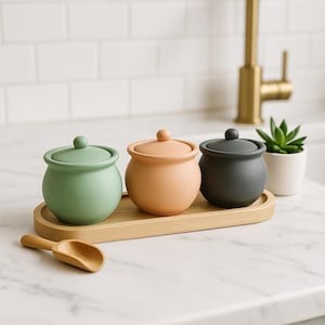 May include: Three ceramic jars in green, peach, and charcoal gray with matching lids sit on a wooden tray. A small wooden scoop rests nearby. A small succulent plant in a white pot is also visible.