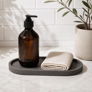 May include: An amber glass soap dispenser with a black pump mechanism, paired with a folded beige waffle-textured cloth, both resting on a smooth, oval-shaped dark grey tray. The items are arranged together on a flat surface.