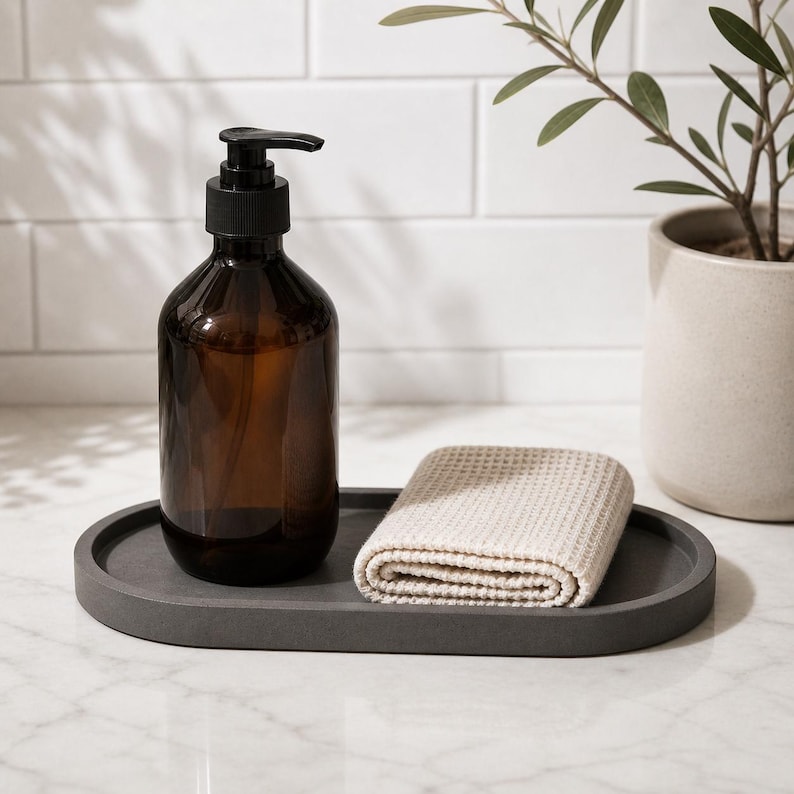 May include: An amber glass soap dispenser with a black pump mechanism, paired with a folded beige waffle-textured cloth, both resting on a smooth, oval-shaped dark grey tray. The items are arranged together on a flat surface.