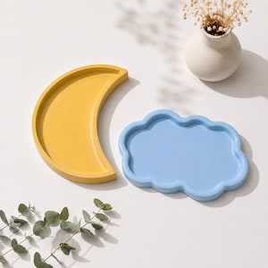 May include: A yellow crescent moon-shaped tray and a light blue cloud-shaped tray. Both trays feature a smooth, matte finish with raised edges, designed as decorative catch-all dishes for small items, presented on a plain, neutral-colored surface.