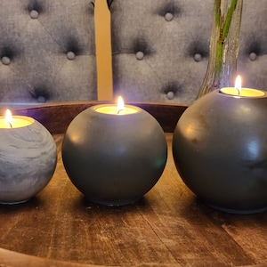 May include: Three spherical candle holders with lit candles. The holders are in shades of gray and sit on a wooden tray. The candles have a warm glow, creating a cozy ambiance. The smallest holder has a marbled effect.