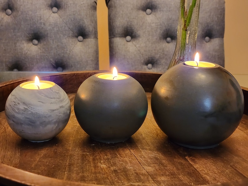 May include: Three spherical candle holders with lit candles. The holders are in shades of gray and sit on a wooden tray. The candles have a warm glow, creating a cozy ambiance. The smallest holder has a marbled effect.