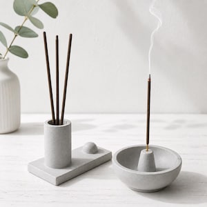 May include: A gray concrete incense holder set. The set includes a cylindrical holder with incense sticks, a rectangular base with a small sphere, and a bowl-shaped holder with a lit incense stick emitting smoke. A white vase with greenery is in the background.