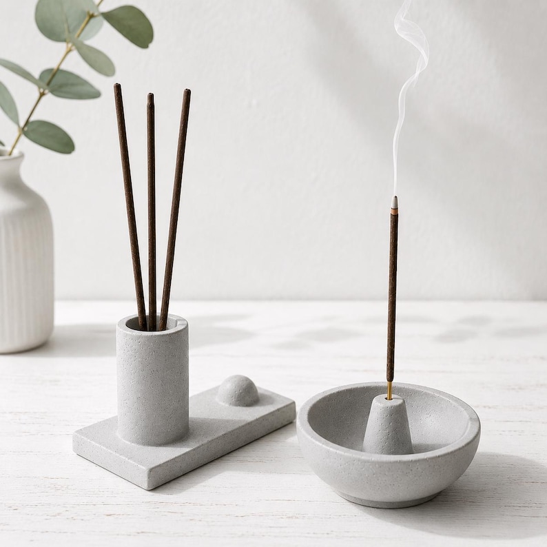 May include: A gray concrete incense holder set. The set includes a cylindrical holder with incense sticks, a rectangular base with a small sphere, and a bowl-shaped holder with a lit incense stick emitting smoke. A white vase with greenery is in the background.