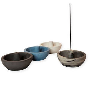 May include: Four stone incense holders in shades of gray, blue, and off-white. Each bowl-shaped holder has a unique design, with one holding a lit incense stick. The holders are arranged on a white background.