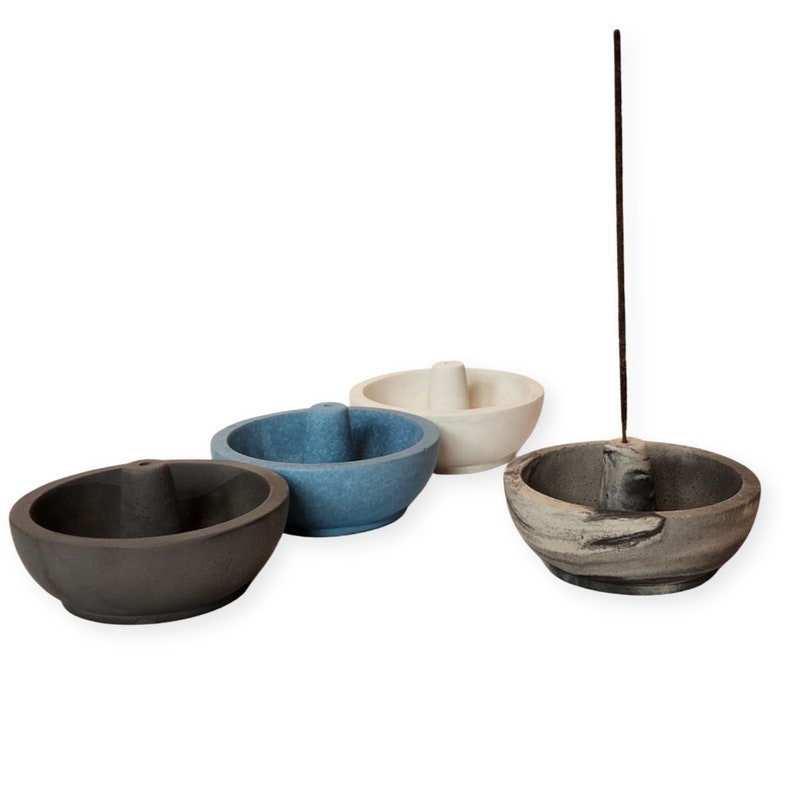 May include: Four stone incense holders in shades of gray, blue, and off-white. Each bowl-shaped holder has a unique design, with one holding a lit incense stick. The holders are arranged on a white background.