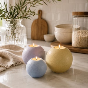 May include: Three spherical candles in pastel colors: lavender, blue, and yellow. Each candle has a lit, small, round candle inside. The candles are on a marble countertop, with a vase of greenery and kitchenware in the background.