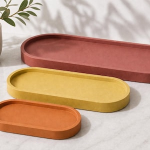 May include: Three oval-shaped trays in varying sizes and colours. The largest tray is a deep terracotta red, the medium tray is a mustard yellow, and the smallest tray is a burnt orange. Each tray features a smooth, matte finish and raised edges.