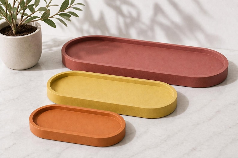 May include: Three oval-shaped trays in varying sizes and colours. The largest tray is a deep terracotta red, the medium tray is a mustard yellow, and the smallest tray is a burnt orange. Each tray features a smooth, matte finish and raised edges.