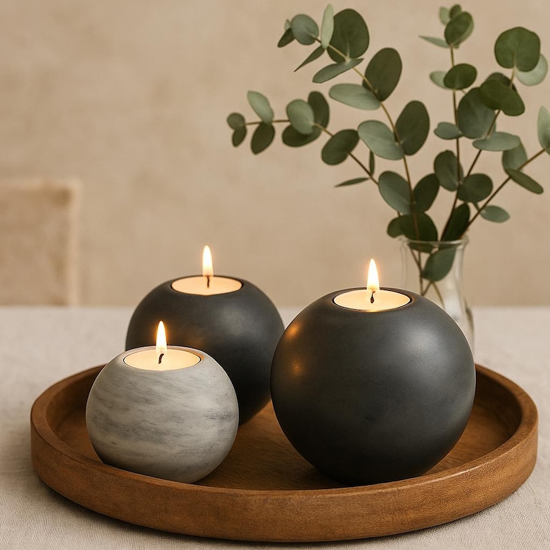 May include: Three spherical candle holders with lit candles sit on a wooden tray. Two are dark gray, and one is a marbled gray and white. A vase of green eucalyptus leaves is in the background, adding a natural element to the scene.