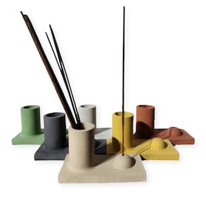 May include: A collection of colorful concrete incense holders. Each holder features a cylindrical design with a square base, available in various colors including green, gray, white, yellow, and terracotta. Incense sticks are placed in the holders.