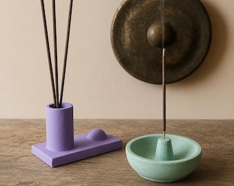 May include: A purple incense holder with three incense sticks and a light green incense holder. A bronze gong hangs in the background. The incense is lit, with smoke rising from it. The objects are on a wooden surface.