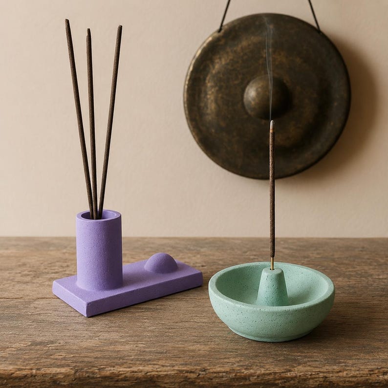 May include: A purple incense holder with three incense sticks and a light green incense holder. A bronze gong hangs in the background. The incense is lit, with smoke rising from it. The objects are on a wooden surface.