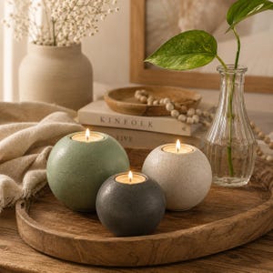 May include: Three spherical candle holders in shades of green, gray, and white, each with a lit candle. They sit on a wooden tray with a vase of greenery and a vase of white flowers in the background. The candle holders are a decorative home accent.