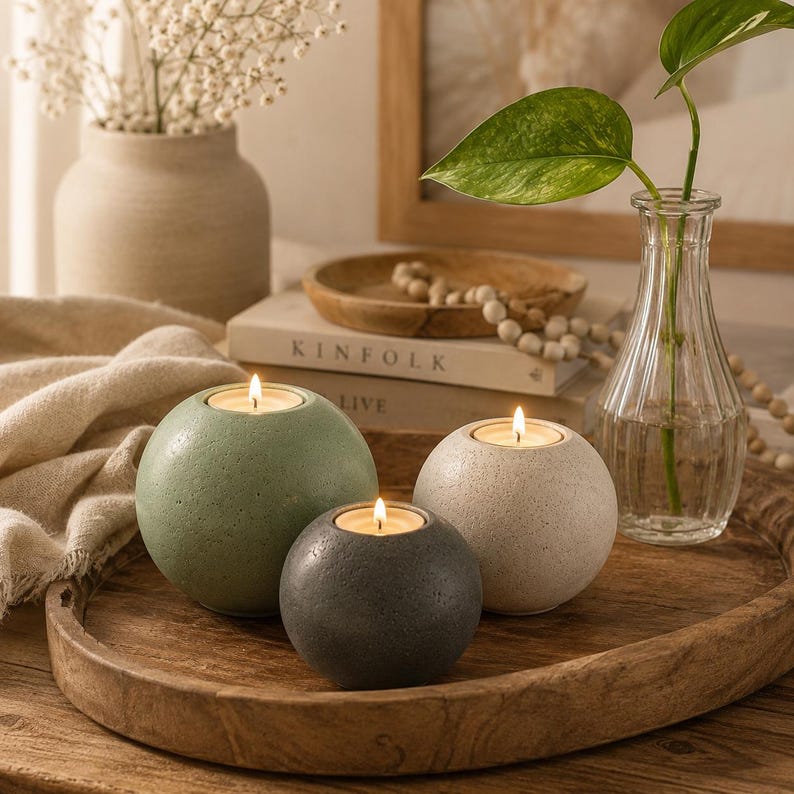 May include: Three spherical candle holders in shades of green, gray, and white, each with a lit candle. They sit on a wooden tray with a vase of greenery and a vase of white flowers in the background. The candle holders are a decorative home accent.