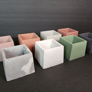 May include: A collection of eight square concrete planters in various colors, including pink, peach, white, green, and gray. The planters are arranged on a dark surface, showcasing their minimalist design and textured finish. Ideal for small plants or succulents.