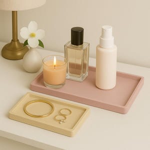 May include: Two rectangular trays, one beige and one pink, with various items on top. The beige tray has a gold bangle and two gold rings. The pink tray has a clear glass bottle of perfume, a white bottle of lotion, and a clear glass candle.