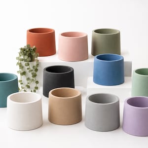 May include: A collection of cylindrical planters in various colors, including terracotta, pink, green, blue, black, white, tan, gray, and lavender. One planter holds a small plant with trailing vines. The planters are arranged on a white surface.