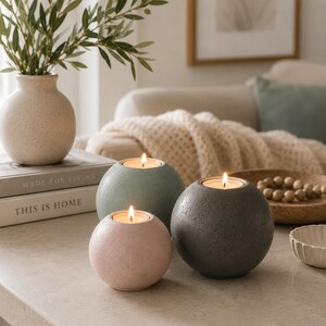 May include: Three spherical candle holders in shades of pink, green, and gray, each with a lit candle inside. A vase with greenery and books with the text "MADE FOR LIVING" and "THIS IS HOME" are in the background. A decorative bowl and beads are also visible.