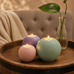 May include: Three spherical candle holders in pastel colors: pink, purple, and green. Each holder has a lit candle inside, creating a warm glow. The holders are on a wooden tray, with a vase of greenery in the background.