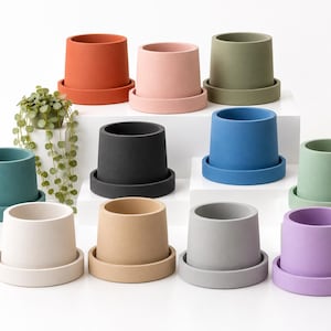 May include: A collection of eleven cylindrical plant pots with matching saucers in various solid matte colors including terracotta, pink, olive green, teal, black, blue, sage, cream, beige, gray, and purple, with a small trailing green plant.