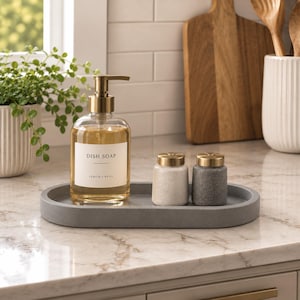 May include: A clear glass dish soap dispenser with a gold pump and a white label reading DISH SOAP LEMON + BASIL, accompanied by two small speckled shakers with gold lids, all arranged on an oval gray concrete tray on a marble countertop.