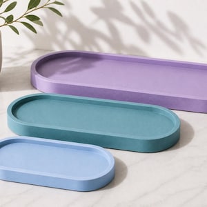 May include: Three oval-shaped trays in graduated sizes, featuring a smooth matte finish. The largest tray is purple, the medium tray is teal, and the smallest tray is light blue. Each tray has a raised rim and a flat, recessed base.