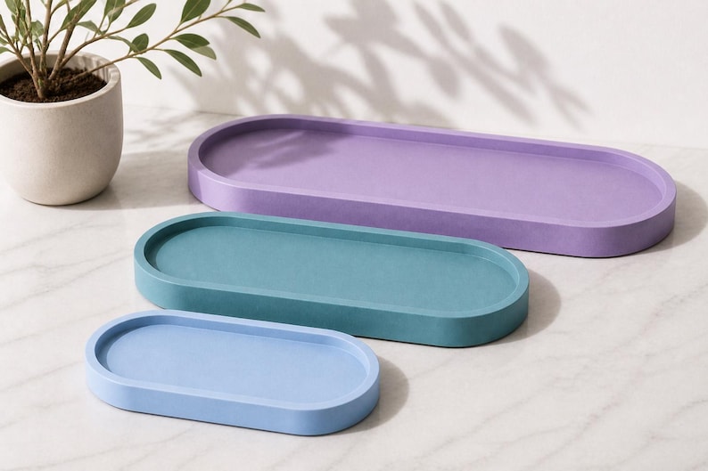 May include: Three oval-shaped trays in graduated sizes, featuring a smooth matte finish. The largest tray is purple, the medium tray is teal, and the smallest tray is light blue. Each tray has a raised rim and a flat, recessed base.