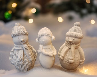 Puede incluir: Tres figuras de muñeco de nieve de cerámica blanca con gorros y bufandas de punto. Los muñecos de nieve están de pie sobre una superficie blanca con un fondo borroso de luces.