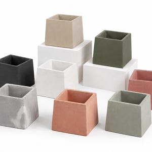 May include: A collection of square, concrete planters in various colors, including black, white, gray, beige, olive green, and coral. The planters are arranged on white platforms, showcasing their geometric design and textured surfaces.