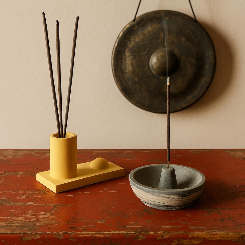 May include: A yellow incense holder with three incense sticks and a small, round, yellow accent piece. A gray and white incense holder with a lit incense stick is in front. A bronze gong hangs in the background.