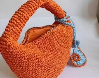 Könnte beinhalten: Eine orangefarbene Häkeltasche mit gebogenem Griff und Reißverschluss. Der Innenraum ist orange. Ein blau-oranger Spiralakzent hängt an der Seite. Die Tasche ist handgefertigt und hat ein strukturiertes Aussehen.