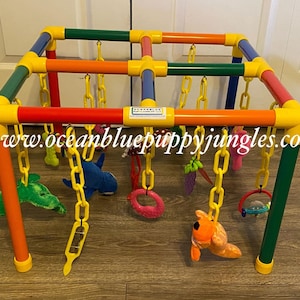 May include: A colorful, multi-level activity center for puppies, made of PVC pipe and featuring hanging toys with chains. The toys include a plush shark, a green plush leaf, a pink ring, a plush dog, a rattle, and a grape bunch. The center is labeled "Oceanblue Puppy Jingles" and includes the website "www.oceanbluepuppyjingles.com".