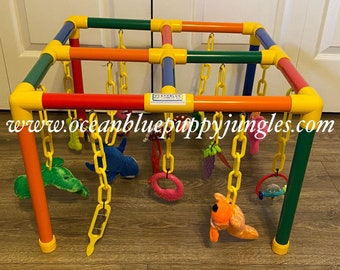 May include: A colorful, multi-level activity center for puppies, made of PVC pipe and featuring hanging toys with chains. The toys include a plush shark, a green plush leaf, a pink ring, a plush dog, a rattle, and a grape bunch. The center is labeled "Oceanblue Puppy Jingles" and includes the website "www.oceanbluepuppyjingles.com".