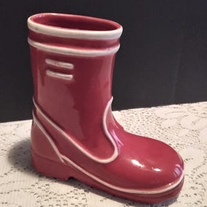 May include: A red ceramic boot shaped vase with white trim. The boot has a glossy finish.