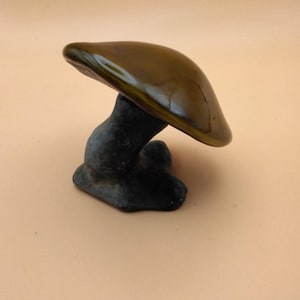 May include: A decorative mushroom figurine featuring a smooth, glossy, dark olive-green cap and a textured, matte black stem. The object has a curved, organic shape with a wide, rounded top that extends over the base, showcasing a contrast in finish.