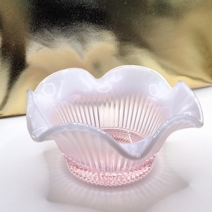 May include: A small, ruffled-edge bowl in soft pink glass. The bowl features vertical ribbed detailing on the interior and a decorative, textured base. The rim is a translucent white, creating a delicate contrast. The bowl is likely vintage or antique.