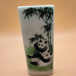 May include: A white rectangular ceramic vase with a hand-painted design of two pandas eating bamboo. The pandas are black and white, and the bamboo is green. The vase has a green base and a slightly tapered shape.