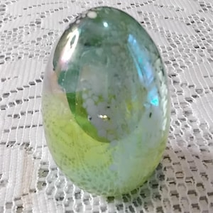 May include: A decorative glass egg with a pearlescent finish. The egg is primarily green with areas of yellow and white. The surface has a textured appearance. The egg is sitting on a white lace tablecloth.