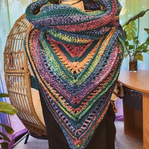 May include: A colorful crocheted shawl with a green, blue, purple, orange, and pink color scheme. The shawl is folded and draped over a mannequin.