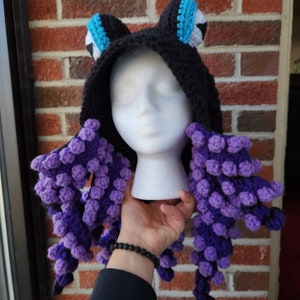 May include: A black crocheted hat with purple and blue crocheted tentacles. The hat has a hood and large blue eyes.