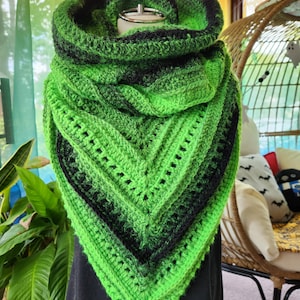May include: A handmade crochet shawl in shades of green and black. The triangular shawl features a gradient effect, transitioning from bright green to black. The cowl neck is also green and black. The shawl is displayed on a mannequin.