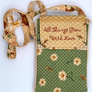 May include: A floral crossbody bag with a daisy and dragonfly pattern. The bag features a green base with white daisies and brown dragonflies, and a tan flap with the embroidered text "All Things Grow With Love". The strap has a daisy pattern.